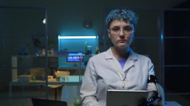 Beyaz önlüklü, gözlüklü, protez kollu, mavi kıvırcık saçlı, kimya laboratuvarında tablet bilgisayarda daktilo eden kendinden emin genç beyaz bir kadının orta boy portresi.