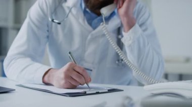 Kafkasyalı erkek pratisyen hekimin hastanedeki hastayla telefonda konuşurken tıbbi geçmişine bilgi yazarken orta eğim görüntüsü.