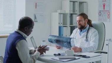 Afrika kökenli Amerikalı hastaları dinleyen ve hastanedeki randevusu sırasında röntgen görüntülerini analiz eden beyaz erkek doktorun orta boy fotoğrafı.