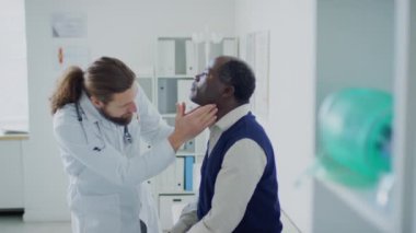 Kafkas hekiminin özel bir klinikte kontrol sırasında kanepede oturan Afrikalı Amerikalı bir hastanın boynundaki tiroid bezlerini kontrol ederken orta boy fotoğrafı.