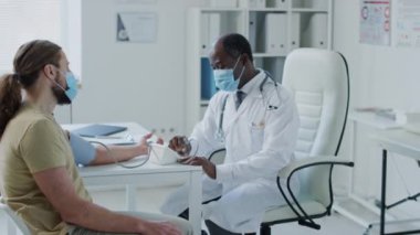 Siyahi erkek doktorun yüz maskesi takıp klinikte sağlık kontrolü sırasında tonometre kullanarak beyaz hastaların sağlığını kontrol ettiği orta ölçekli bir fotoğraf.