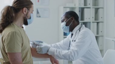 Afrika kökenli Amerikalı bir doktorun yüz maskesi ve eldiven taktığı ve klinikte yüz maskesi takan beyaz bir hastaya aşı yaptığı orta boy bir fotoğraf.