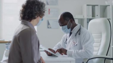 Yüz maskesi takan kadın hastanın yüz maskesi takan ve elinde tablet olan siyah erkek doktora şikâyet ettiği orta boy bir fotoğraf.