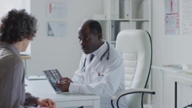 Afro-Amerikalı doktorun tablet tutarken orta boy fotoğrafı ve hastanedeki beyaz kadın hastaya MRI sonuçlarını açıklarken.