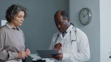 Beyaz kadın hastanın yanında duran Afrikalı Amerikalı doktorun orta boy fotoğrafı ve klinikteki tıbbi kayıtları gösteriyor ki...