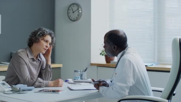 白种人女性患者双手抱头 向黑人男性医生讲述在医院预约时头痛破裂的中侧照像 — 图库视频影像