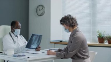 Yüz maskeli siyah erkek doktorun, klinikteki randevusu sırasında yüz maskesi takan beyaz kadın hastaya göğüs röntgeni sonuçlarını gösterip açıkladığı orta ölçekli bir fotoğraf.