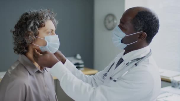 顔面マスクでアフリカ系アメリカ人医師のメディアショット 顔面マスクを着用している女性の白人女性の首に甲状腺をチェックし 病院で医療記録に結果を書く — ストック動画