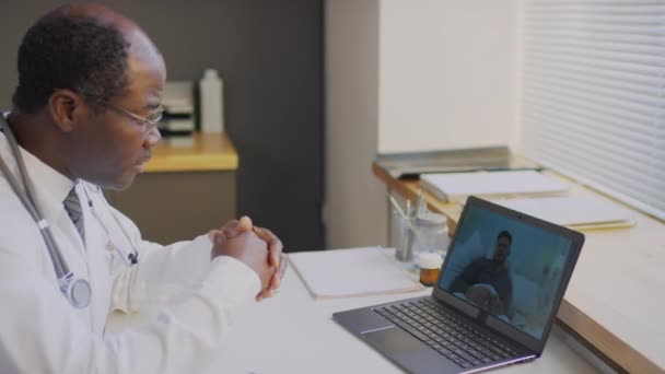 ラップトップ上のオンラインビデオコールを介して病院に滞在する患者をコンサルティングする黒人男性開業医の中側ショット — ストック動画