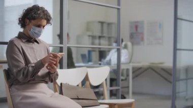 Kafkasyalı bir kadının hastane koridorunda sırasını beklerken yüz maskesi takıp telefon kullandığı orta boy bir fotoğraf.
