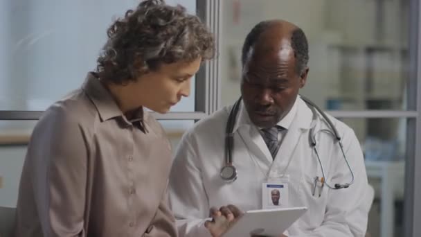 白种人女性患者与黑人男性医生坐在医院走廊中 聆听平板屏幕上的诊断说明 — 图库视频影像