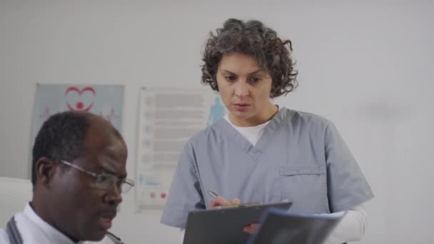 メディアサイドハンドヘルドショット黒人男性経験豊富な医師が胸部X線スキャンを保持し 診療所の医療記録で情報を書く女性の白人同僚に診断を説明 — ストック動画