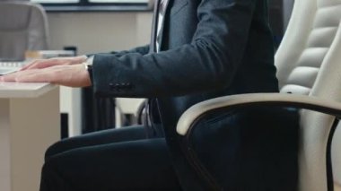 Tanımlanamayan takım elbiseli bir ofis çalışanının, bilgisayar sandalyesinde masa başına doğru ilerlerken ve dizüstü bilgisayarla çalışmaya devam ederken görüntüsü.