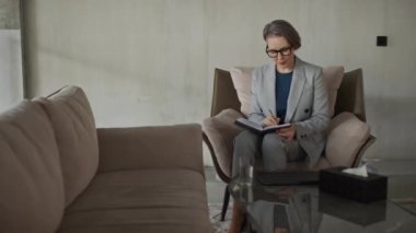 Koltuğunda oturan kadın psikoloğun modern ofisteki nötr renkli iç mekan ile ilgili notlar aldığı bir fotoğraf.