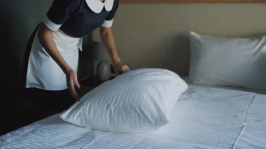 Siyah beyaz üniformalı hizmetçinin rahat otel odasında yastık kabartma ve battaniye düzeltirken orta boy bir fotoğrafı.