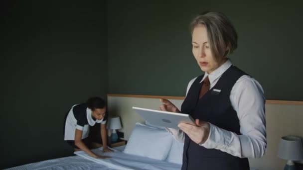 女性ホテルのマネージャーがベッドを作っている間にメイドをチェックするための選択的なフォーカス媒体ショット — ストック動画