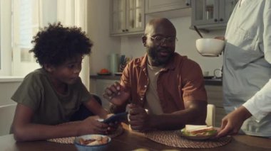 Orta boy Afro-Amerikan bir çocuk ve babası. Mutfak masasında birlikte oturuyorlar, akıllı telefonlara bakıyorlar. Hemşire üniformalı anne sandviç, mısır gevreği ve süt koyuyor.