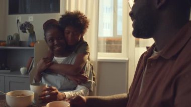 Orta boy Afro-Amerikan sağlık çalışanı üniformalı evde oturmuş, kahvaltı yapıyor, kahve içiyor, küçük oğlu annesine sarılıyor ve kocasıyla sohbet ediyor.