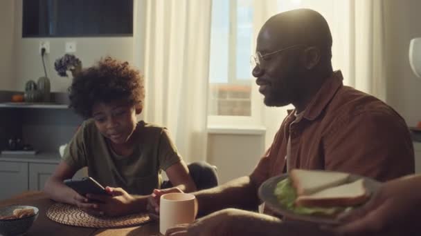 若い黒人の少年と父親がキッチンで一緒に座って スマートフォンでゲームをしたり チャットしたり 医療ユニフォームで母親がシリアルのボウルを持ってきてミルクを注ぐ中型ショット — ストック動画