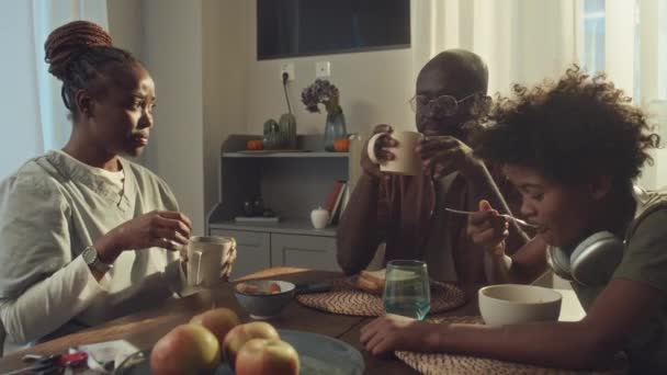 身着医疗制服的年轻非洲裔美国妇女与丈夫和儿子坐在厨房餐桌旁 吃早饭 然后看表 拿钥匙 说再见 赶忙去工作 拍了一张中等的照片 — 图库视频影像