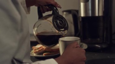 Hastane üniformalı siyahi kadın sıhhiyecinin evde mutfakta durup bardaktan bardağa kahve dökerken ve içerken mesaiden önce sandviç yerken yakın çekimde çekilmiş fotoğrafı.