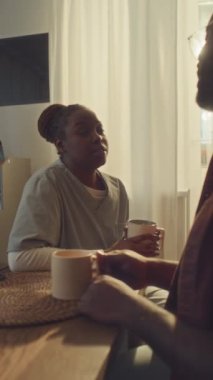 Orta boy dikey fotoğraf, mercekli genç Afro-Amerikan kadın hemşire üniforması ile mutfakta kocasıyla oturuyor, çay içiyor, sohbet ediyor ve iş problemlerini tartışıyor.