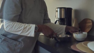 Kapalı çekimde, hastane üniformalı Afro-Amerikan kadının evde mutfakta durup, kaseye evcil hayvan maması koyarken, köpek yemesi için yere koyarken ve okşarken.