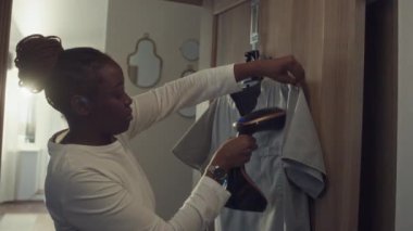 Afro-Amerikalı kadın sıhhiyecinin evde koridorda dikey el yapımı buharlı iş üniformasını ütülerken, vardiyaya hazırlanırken orta boy fotoğrafı.