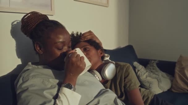 病院でアフリカ系アメリカ人女性看護師のミディアムクローズアップショット シフトする前に10代の息子と家でソファーでリラックスして 抱擁し コーヒーを飲んで 男の子の髪とチャット — ストック動画