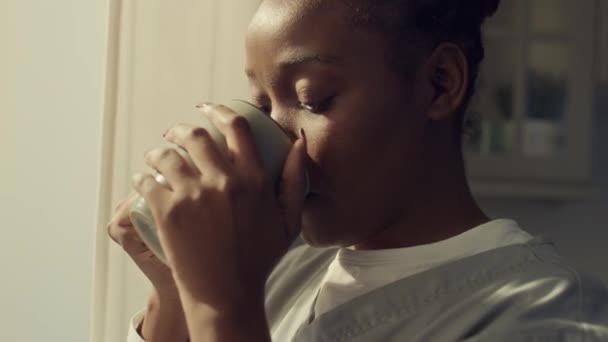 身着卫生工作者制服的美丽而沉思的非洲裔美国妇女站在窗边 凝视着窗外 沉思着什么 喝着杯子里的咖啡 拍了一张特写镜头 — 图库视频影像