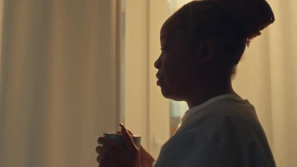 身着医院制服 头脑清醒的年轻非洲裔美国医务工作者走向窗户 凝视着外面 喝咖啡 然后在黎明时分提早下班 这对年轻的非洲裔美国医务工作者进行了跟踪调查 — 图库视频影像