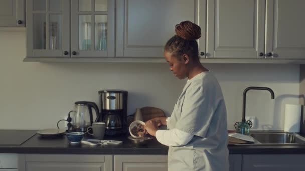 自宅でキッチンに立っている健康労働者の制服の黒人女性のメディア傾斜ショット ペットフードをボウルに入れ マルチポーのために床に置き ストロークし 犬が食べる — ストック動画