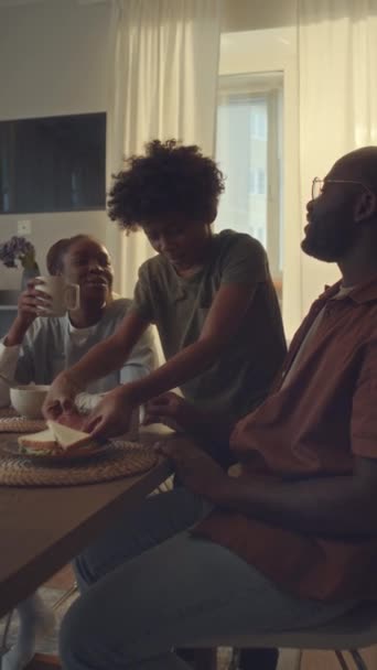身着制服的非洲裔美国女医务工作者在工作前与家人共进早餐的中垂直镜头 年幼的儿子抱着爸爸 吃三明治 聊天和开玩笑 都面带微笑 — 图库视频影像