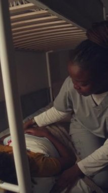 Orta boy, Afro-Amerikan bir çocuğun gece yatakta uyuduğu, hastane üniformalı bir annenin içeri girdiği, yorganı içeri soktuğu, onu yanağından öptüğü, sonra da yatak odasının kapısını kapattığı dikey bir fotoğraf.