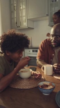 Orta boy Afro-Amerikan çocuk ve babasının mutfakta birlikte otururken, kahvaltı ederken, hemşire üniforması içinde mısır gevreğine süt dökerken, sohbet ederken ve gülümserken orta boy dikey pozu.