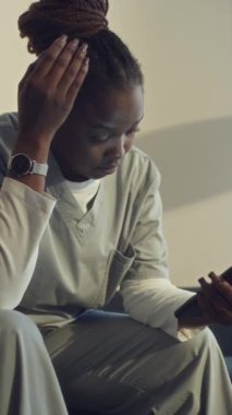 Genç Afrikalı Amerikalı doktorun işten sonra evde oturup, akıllı telefondaki vardiya çizelgesine bakarken, başını sallarken ve boyun kaslarına masaj yaparken orta boy dikey pozu.