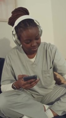 Medium dikey çekimde, tıbbi üniformalı ve kulaklıklı genç siyahi bir kadın bacak bacak bacak üstüne atmış koltukta oturmuş akıllı telefondan müzik dinliyor ve şarkının ritmine göre sallanıyordu.