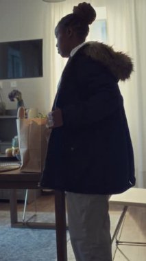 Uzun vardiyadan sonra eve dönen yorgun, siyah kadın hastane çalışanı ve parka ceketinin orta boy dikey pozu. Paket içinde alışveriş poşetleri taşıyor, masaya yaslanıyor ve esniyor.
