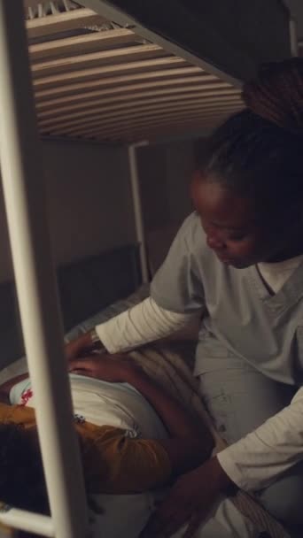 非洲裔美国男孩晚上睡在床上 身穿医院制服的妈妈进来 把羽绒被塞进被窝 亲吻他的脸颊 然后离开并关上卧室大门的中等垂直镜头 — 图库视频影像
