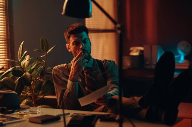 Genç erkek ofis çalışanı sigara içiyor ve iş yerinde karanlık bir ofiste otururken fotoğraflara bakıyor ve işten sonra dinleniyor.