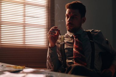 Retro forma giymiş genç düşünceli bir adam karanlık bir ofiste masa başında oturmuş sigara içiyor.