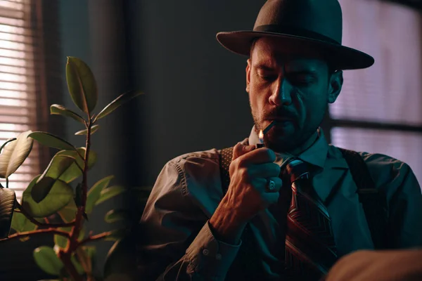 Young retro man in hat and formalwear lighting cigarette while sitting in front of camera with green domestic plant on his right