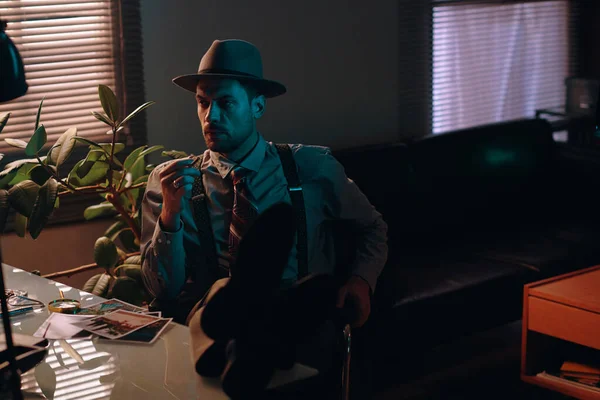 年轻而严肃的商人 头戴礼帽 身穿正装 坐在黑暗的办公室里 一边看一边抽烟 — 图库照片