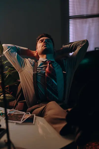Young man in retro clothes smoking cigarette while relaxing by workplace in dark office at break with his hands on back of head