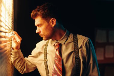 Meraklı genç iş adamlarının eski moda kıyafetleriyle ofiste pencerenin önünde dikilirken jaluziden bakan bakışları.