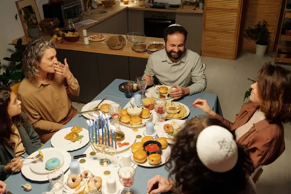 Happy family of five having chat and eating homemade food while sitting by served table and celebrating holiday of Hanukkah