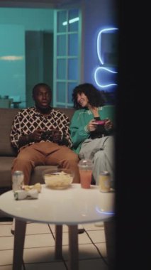 Kanepede oturan ve TV 'de neon ışıklı modern apartmanda oyun oynayan farklı arkadaşların dikey çekimleri.