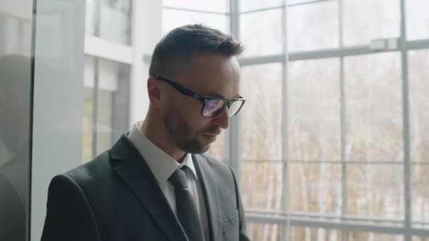 スマートフォンを使ったメガネとグレーのスーツでコーカサス人のビジネスマンのチルトスローモショット 現代オフィスのパノラマエレベーターで下りる — ストック動画