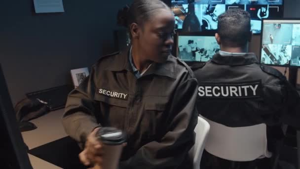 ガードルームの制服の監視Cctvビデオ映像を一緒に制御する男性Biracialの同僚にコーヒーを与える若い黒人女性ガードの中型ハンドヘルドショット — ストック動画