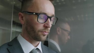 Yakın plan çekimde, beyaz erkek yönetici gözlüklü ve takım elbiseli, panoramik asansörle aşağı iniyor ve modern ofis saatlerinde gözlerini kaçırıyor.